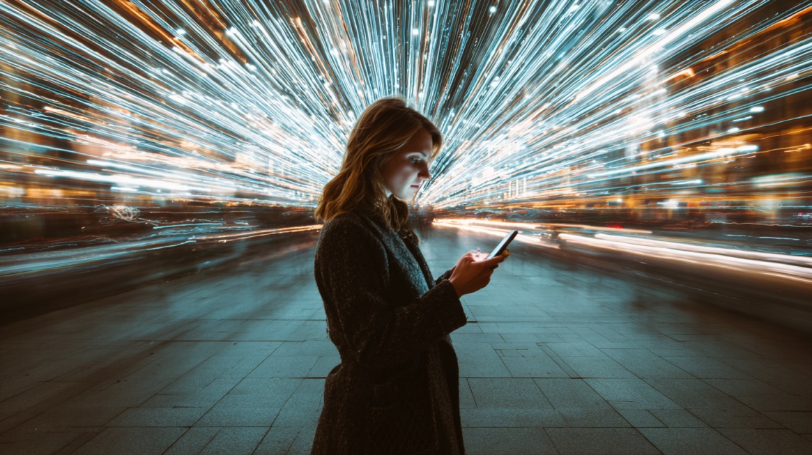 Žena stoji napolju noću i gleda u svoj telefon, dok svetlosne linije u pozadini stvaraju efekat velike brzine i kretanja