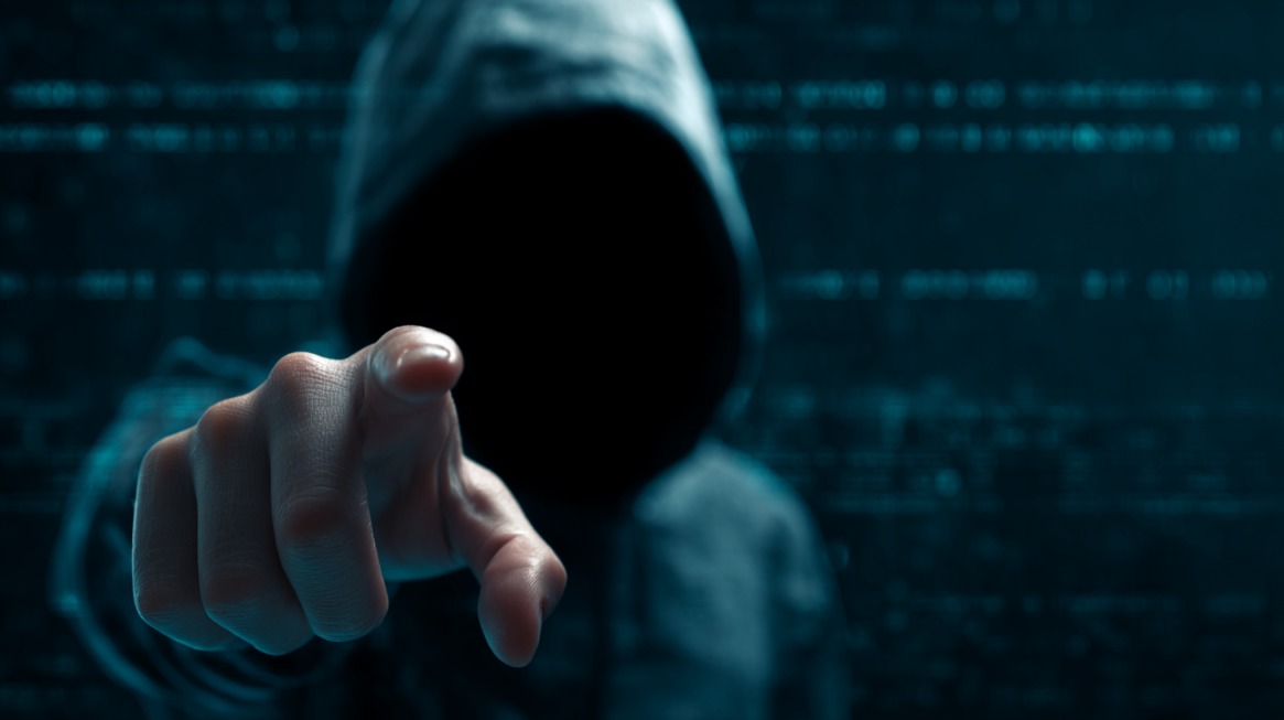 Figura osobe u tamnoj dukserici sa kapuljačom, nevidljivog lica, pruža ruku ka kameri u mračnom okruženju ispunjenom digitalnim kodovima