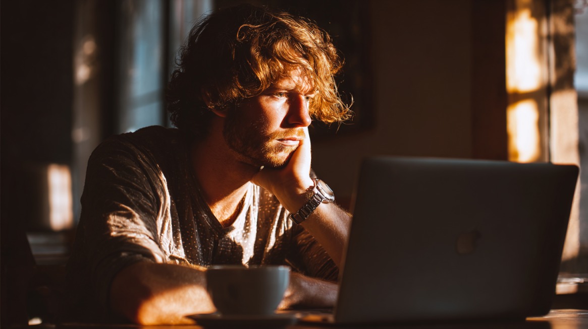 Mladi muškarac sa dugom kosom sedi za laptopom u toplom ambijentu, osvetljen zlatnim svetlom sunca dok koncentrisano radi