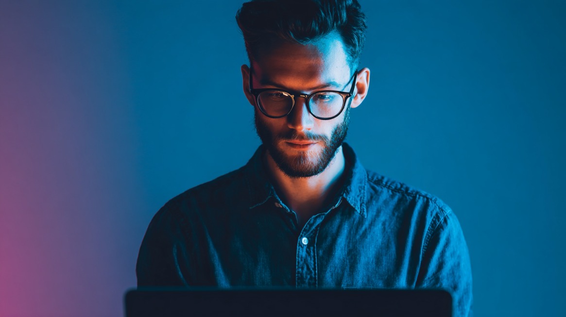 Mladi muškarac sa naočarima sedi ispred laptopa, osvetljen plavim i ružičastim svetlom, fokusirano radići na svom online projektu